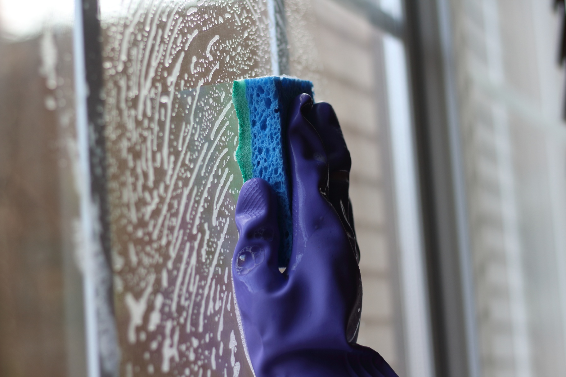 Gloved hand cleaning a window with a soapy sponge